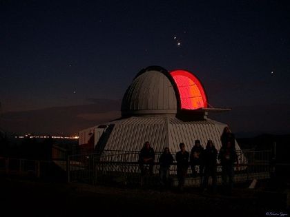 Nighttime photo of a white building with its open dome giving off red light and people observing the star-filled sky