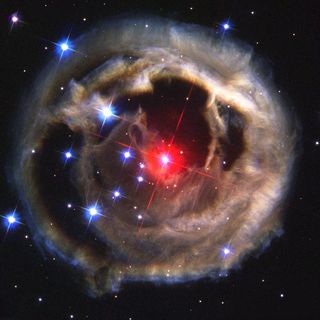 Photo of a very luminous red star surrounded by a mass of beige clouds and luminous white dots against a very black background