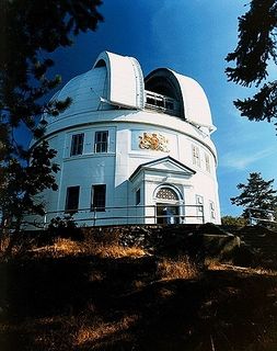 Photo couleur d'un bâtiment blanc à dôme ouvert, situé en haut d'une colline boisée