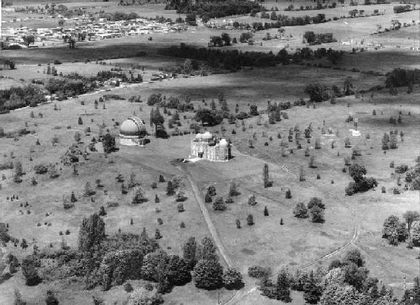 Black and white photo of two buildings with domes, with a scattering of young trees nearby and a residential neighbourhood at top left