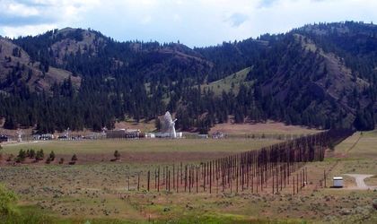 Photo d'une série de poteaux créant un mur dans la plaine verte, à droite d'une antenne blanche entourée de bâtiment et devant des montagnes boisées.