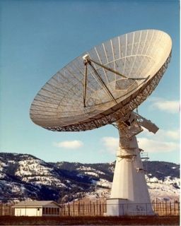 Photo d'une antenne parabolique métallique blanche pointant vers le ciel, clôturée et située à droite d'un petit bâtiment blanc avec, en arrière-plan, des collines enneigées