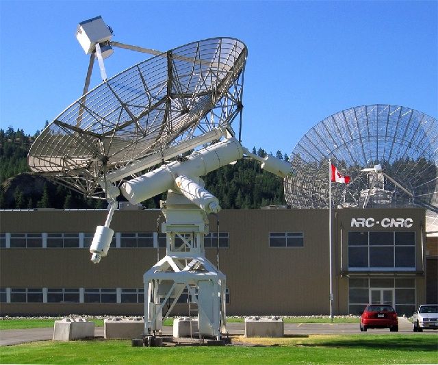 Photo of a white metal antenna pointing towards the sky located in front of a beige building partially hiding another antenna