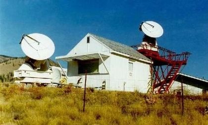 Photo d'un bâtiment rectangulaire blanc et de deux antennes pointant vers le ciel bleu, entouré de hautes herbes verdâtres.