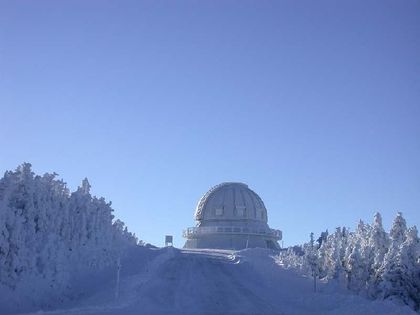 Photo d'un dôme blanc, situé au bout d'un chemin boisé recouvert d'une épaisse couche de neige blanche, devant un ciel bleu