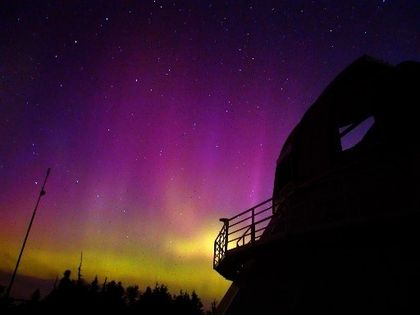 Photo d'un bâtiment à dôme, à contre-jour, devant une aurore boréale aux teintes jaunes et mauves dans un ciel étoilé