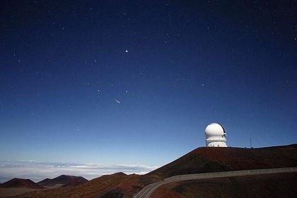 Photo of a landscape with a white observatory to the right located on a barren brown hill under a starry night changing from a white to a midnight blue sky