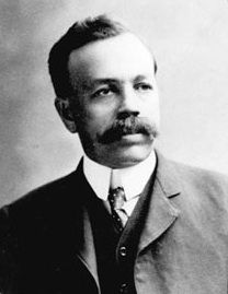 Black and white photo of a man with a large mustache, wearing a suit and tie and looking to his right