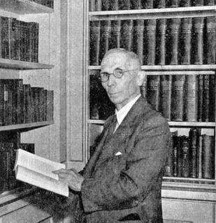 Black and white photo of a man in profile looking at the lens, holding a book in his hands and standing in front of a bookcase