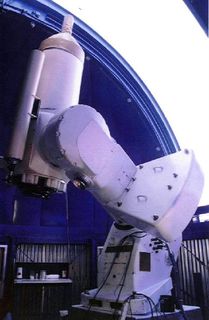 Photo d'un télescope blanc devant un dôme bleu ouvert sur un ciel très blanc