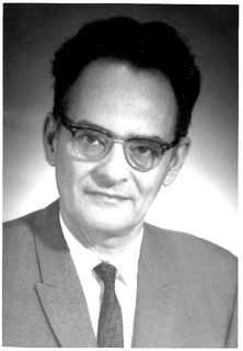 Black and white photo of a man with glasses and short black hair wearing a suit and tie