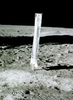 Photo d'une bande de tissu verticale, installée sur le sol lunaire, derrière des traces de pas dans le sol et devant un ciel très noir