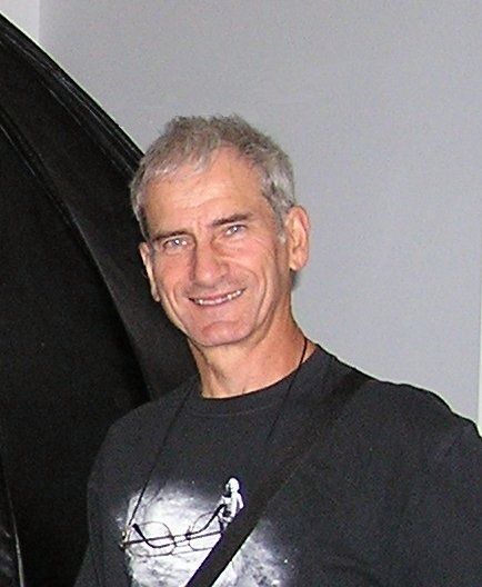 Colour photo of a man with short curly hair wearing a t-shirt and glasses hanging around his neck