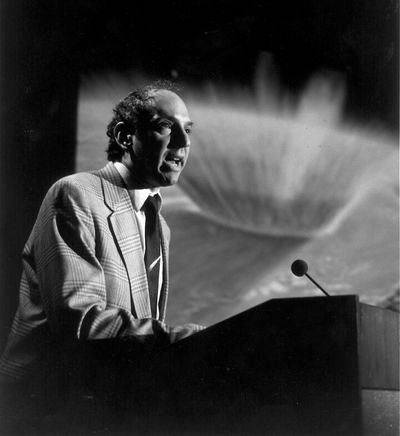 Black and white photo of a man wearing a suit, leaning on a lectern and speaking into a microphone with an explosion projected onscreen in the background