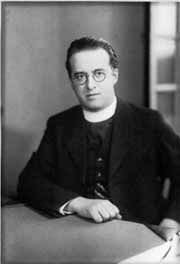 Black and white photo of of a man with glasses, wearing a black jacket and seated at a table