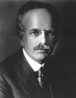 Black and white photo of a man with a mustache looking at the lens, wearing glasses and a suit