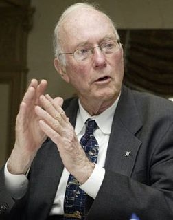 Colour photo of a man with white hair, wearing glasses and a grey suit, gesticulating and speaking at the same time.