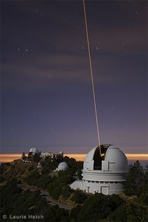 Photo d'un bâtiment blanc avec le dôme ouvert, installé sur une montagne devant un ciel à la pénombre, un faisceau laser jaune pointe vers le ciel