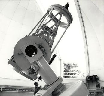 Black and white photo of a grey telescope pointing towards the sky through the opening of a white dome
