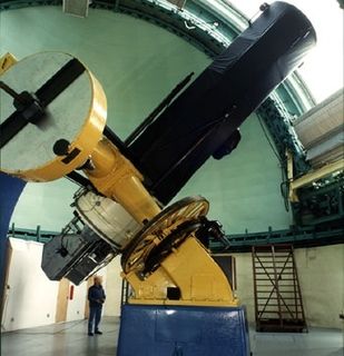 Photo couleur d'un télescope à la base bleue, au corps jaune et au tube télescopique noir pointant l'ouverture d'un dôme blanc
