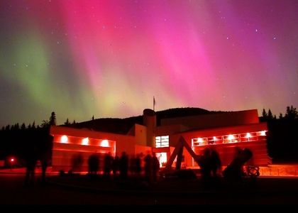Photo nocturne d'un bâtiment illuminé devant une aurore boréale aux teintes vertes et rosées. Des personnes sont debout devant le bâtiment et regardent le ciel.
