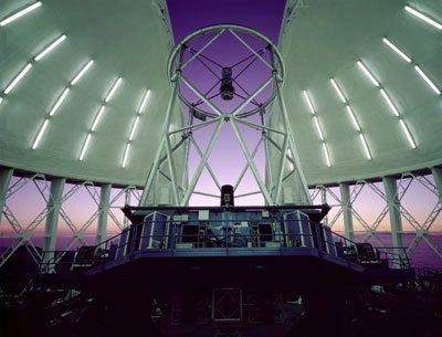 Photo prise de l'intérieur d'un l'observatoire ayant le bas des murs relevés, un dôme blanc ouvert et un télescope pointant vers un ciel mauve au crépuscule