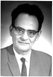Photo noir et blanc d'un homme en complet et cravate, ayant de courts cheveux noirs et portant des lunettes