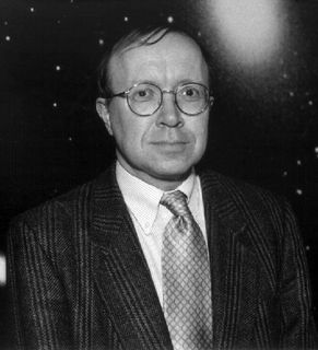 Photo en noir et blanc d'un homme en complet portant des lunettes, devant une image d'un ciel étoilé