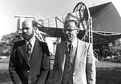 Black and white photo of two men in suits looking to their left and standing in front of a horizontal conical structure