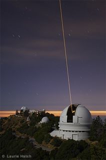 Photo of a white building with the dome open, located on a mountain with a dusky sky in the background and a yellow laser beam pointing towards the sky
